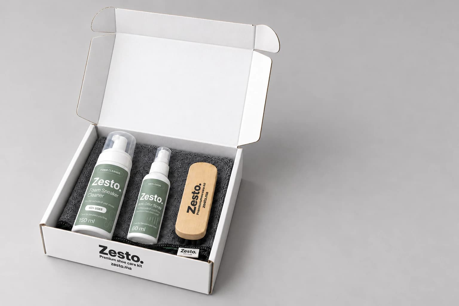 Boîte blanche Zesto. ouverte avec une mousse 150 ml, un spray 60 ml, une brosse en bois et une microfibre grise, sur fond gris clair.